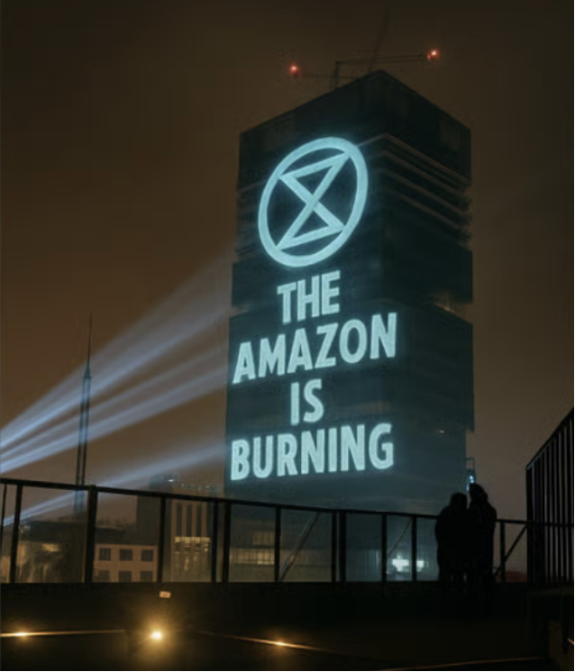 The image is dominated by a building much taller than all the others on which is projected in greenish-white light the XR logo and the words THE AMAZON IS BURNING.