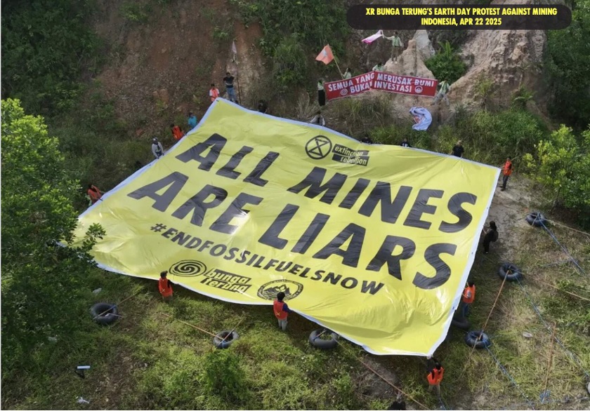 Photo of a large yellow banner held by protesters in a forested area, withtext "ALL MINES ARE LIARS #ENDFOSSILFUELSNOW".