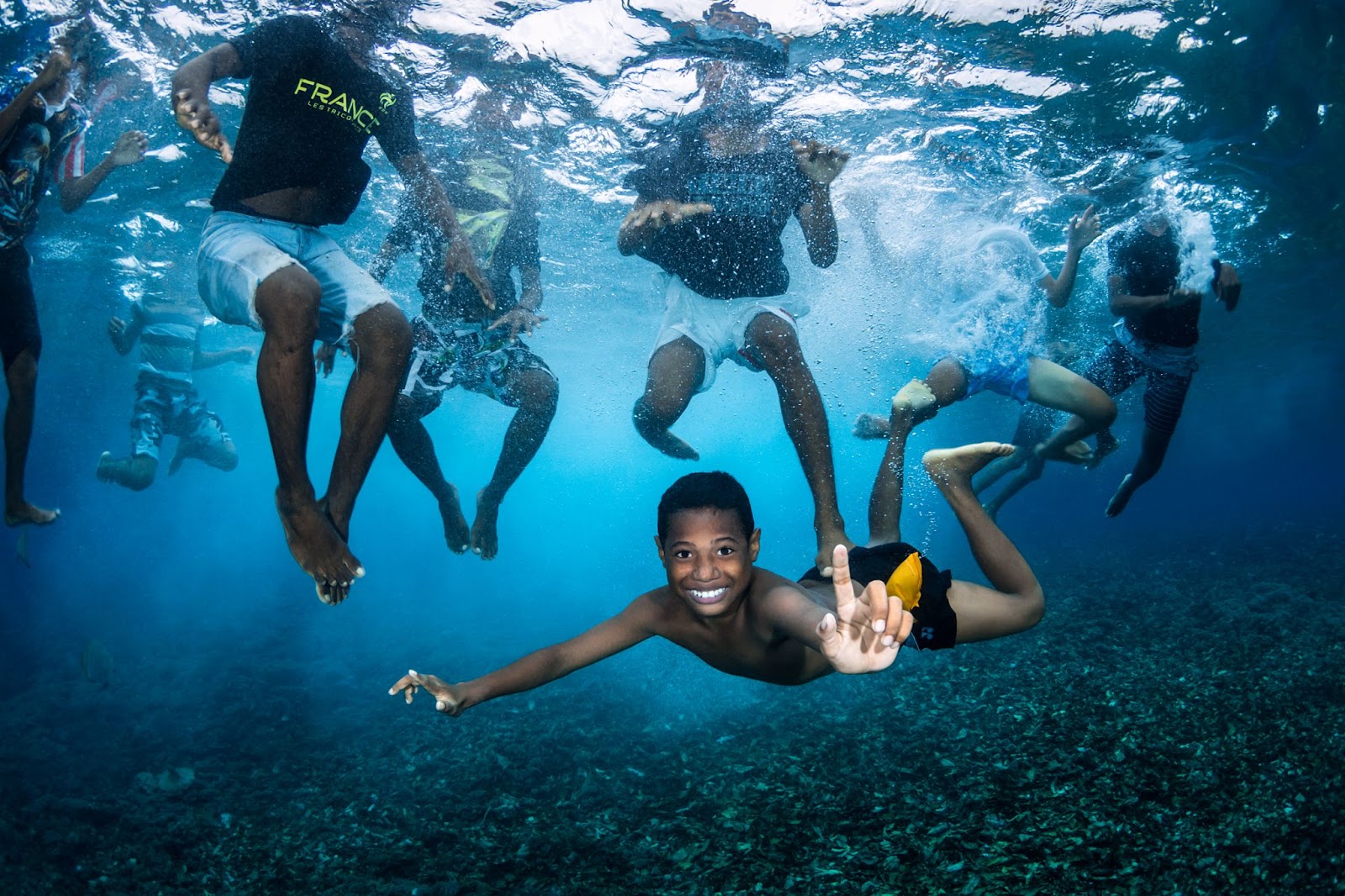 Da sotto l'acqua, vediamo otto ragazzi che nuotano. Sette di loro hanno latesta fuori dall'acqua e non possiamo vederne il volto, ma un ragazzo hanuotato sott'acqua e sta galleggiando orizzontalmente, sorridendo eindicando versol'alto.