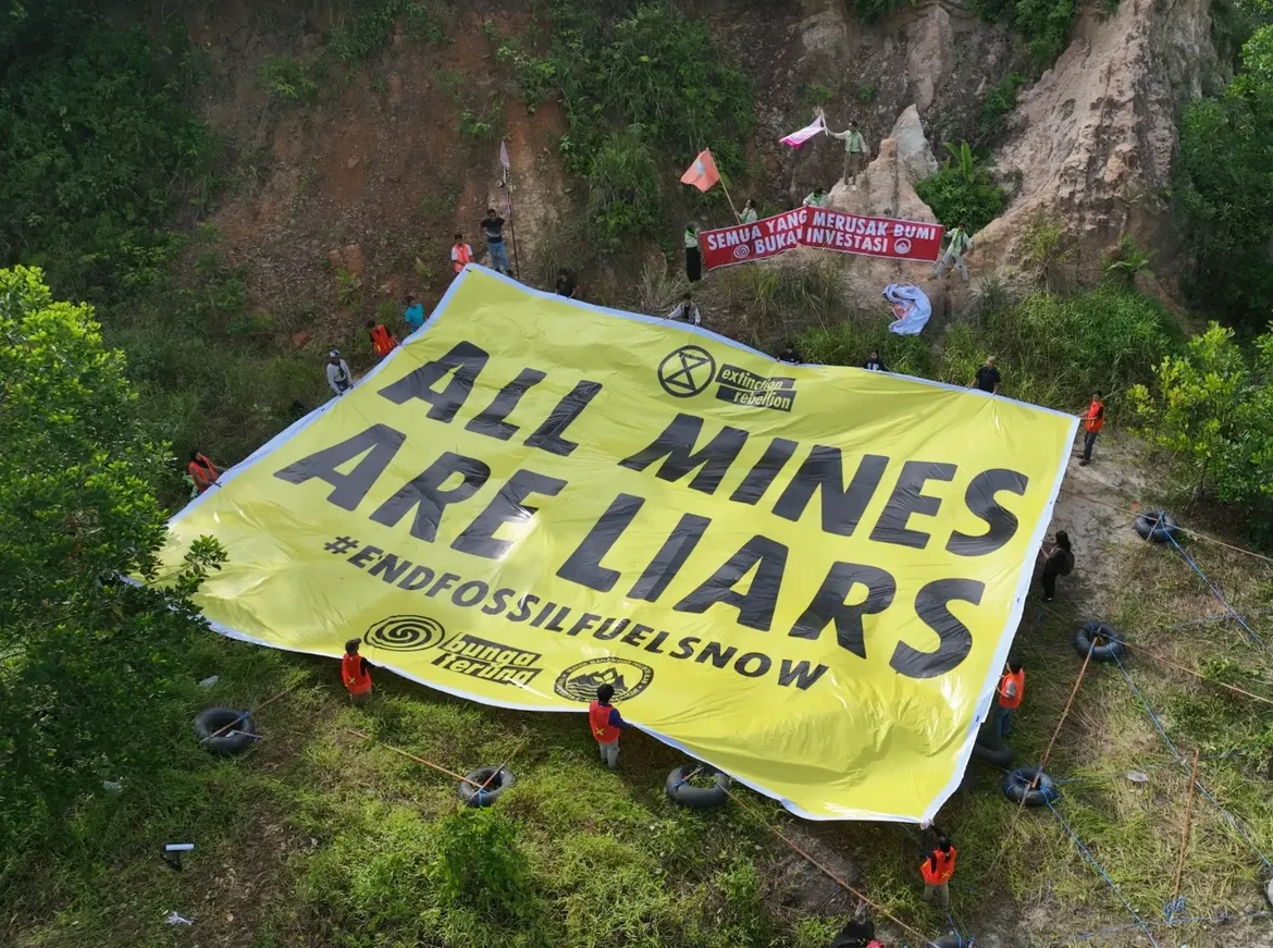 An aerial photo of a group of people outdoors holding a very large sign reading ALL MINES ARE LIARS #ENDFOSSILFUELSNOW